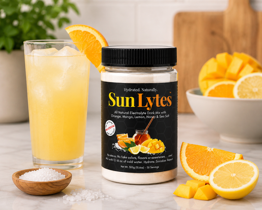 A jar of SunLytes electrolyte drink mix stands on a marble countertop beside a glass of yellow iced drink with an orange slice, with bowls of citrus and salt in a bright kitchen setting.