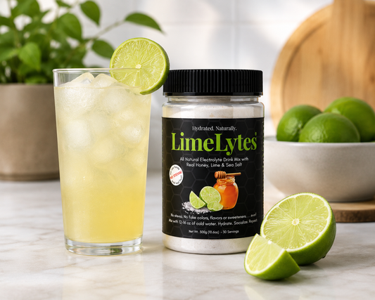 A jar of LimeLytes electrolyte drink mix stands on a marble surface beside a tall glass of pale yellow iced drink with a lime wheel garnish. Limes and a bowl of limes are visible in the softly blurred background, along with greenery and a wooden board.