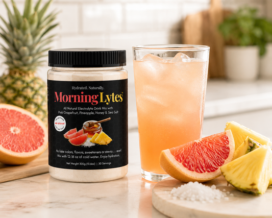 A jar of MorningLytes supplement powder stands on a marble countertop beside a glass of pink iced drink, with sliced grapefruit and pineapple arranged around it in a bright kitchen setting.